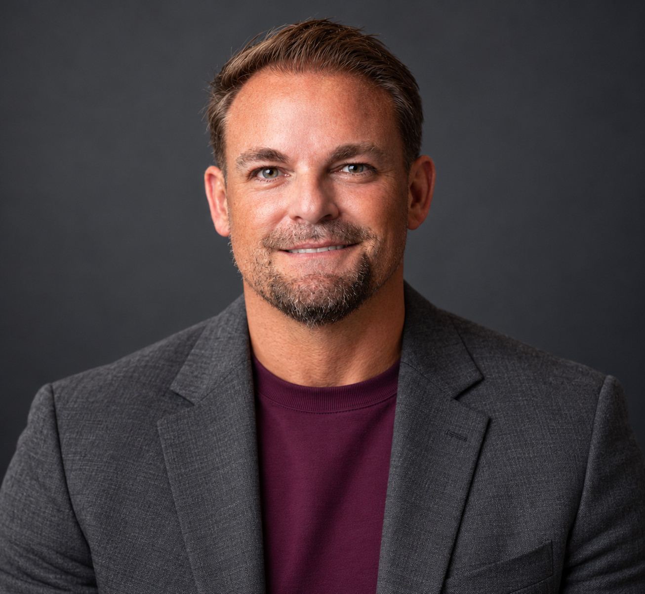 Brent O'Mara smiling in a grey blazer and maroon crew neck against a dark grey studio backdrop