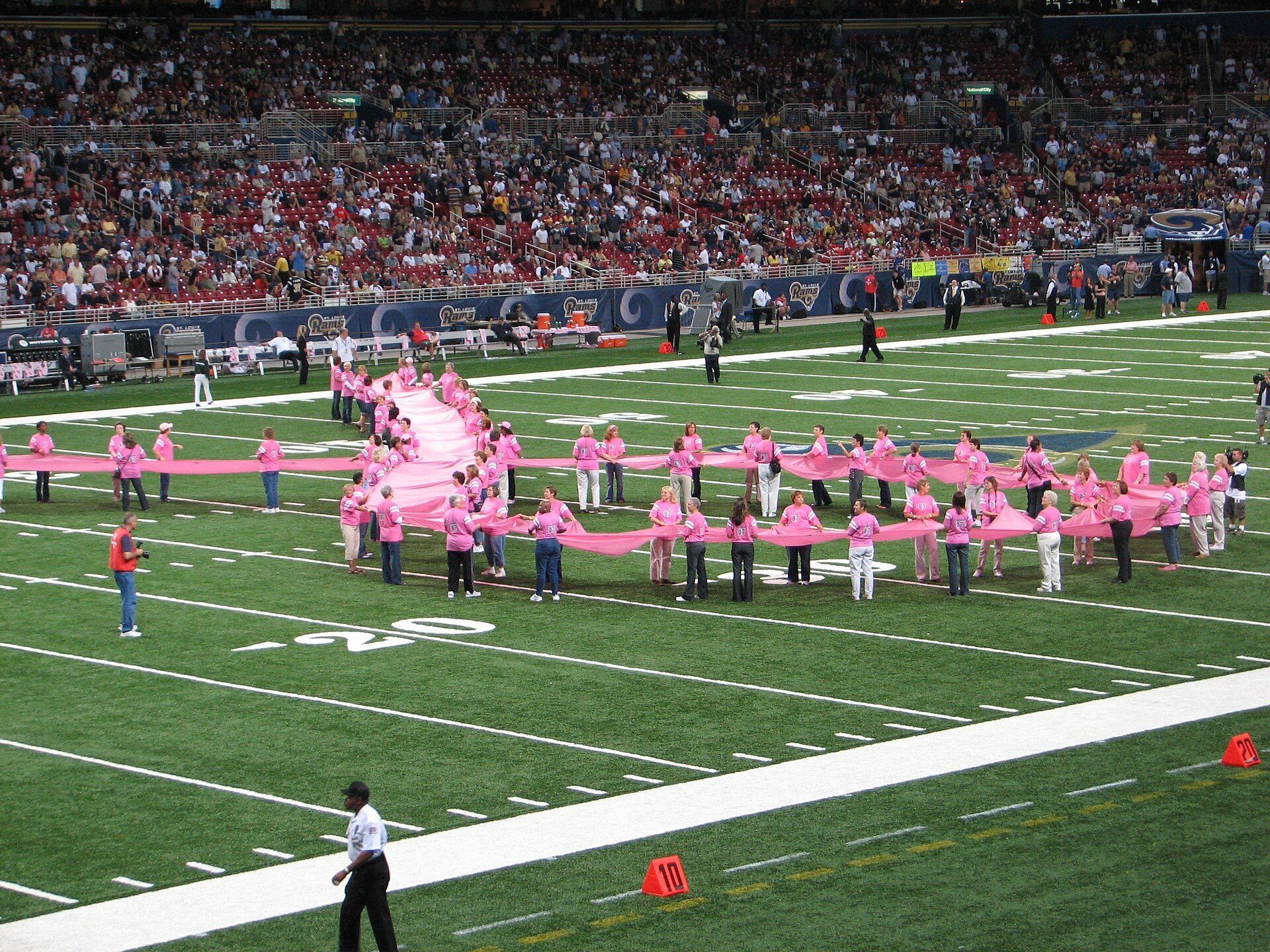 A giant pink ribbon is carried across a football field by volunteers seen from a distance. A Wikimedia Commons photograph on Brent O'Mara Insights.